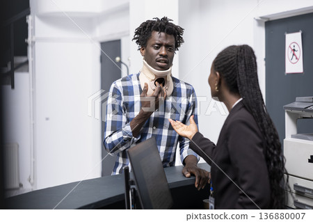 Young black man in neck brace checks with female receptionist to verify doctor appointment. Hospital administrator assists injured male patient, offering medical guidance and support. Young black man in neck brace checks with female receptionist to verify doctor appointment. Hospital administrator assists injured male patient, offering medical guidance and support. 136880007