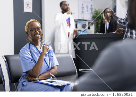 African American female nurse sits in clinic reception area, getting ready for duties and patient care. Cheerful medical professional looks at camera with confidence while waiting in hospital lobby. African American female nurse sits in clinic reception area, getting ready for duties and patient care. Cheerful medical professional looks at camera with confidence while waiting in hospital lobby. 136880021