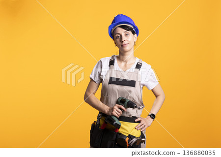 Female adult stands against isolated background wearing construction workwear and safety helmet, holding drill gun with confidence. Female renovator demonstrates home improvement professionalism Female adult stands against isolated background wearing construction workwear and safety helmet, holding drill gun with confidence. Female renovator demonstrates home improvement professionalism 136880035