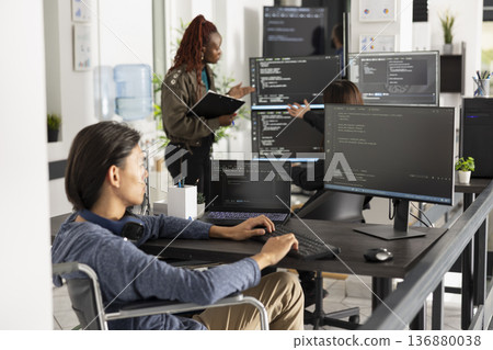 Asian programmer in wheelchair types code at office desk with focused concentration. Software developer analyzes backend data, representing accessibility, inclusion and innovation in technology. 136880038