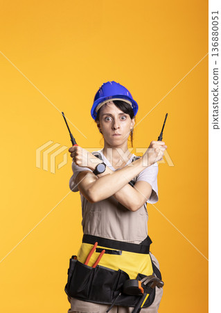 Woman contractor with furious expression, holding construction tools in X shaped arm gesture. Serious female renovator stands in wizardry pose with screwdrivers, determined to tackle building projects 136880051