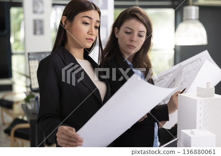 Female architects compare design details and plan execution timelines to ensure smooth project development. Focused women review printed building blueprints in preparation for construction site visit. Female architects compare design details and plan execution timelines to ensure smooth project development. Focused women review printed building blueprints in preparation for construction site visit. 136880061