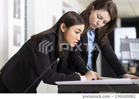 Diverse female professionals working on building designs and detailed floor plans for upcoming projects. Focused women architects collaborate closely while reviewing detailed blueprints in office. 136880062