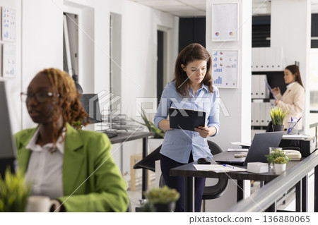 Determined businesswoman stands at desk, comparing data on clipboard with digital reports on laptop. Corporate advisor reviewing analytics, project updates and company performance details. 136880065