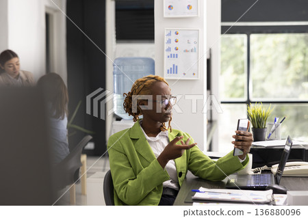 Black project manager sits with smartphone in hand, discussing with client on video call. Professional female employee with earbud, using mobile device for virtual meeting in corporate office. 136880096