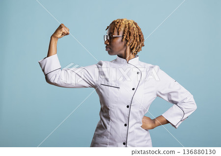 Professional chef looking at flexed arm and expressing empowerment and strength in culinary profession. Black woman raises arm proudly, embracing power, capability and dedication in cooking journey. Professional chef looking at flexed arm and expressing empowerment and strength in culinary profession. Black woman raises arm proudly, embracing power, capability and dedication in cooking journey. 136880130