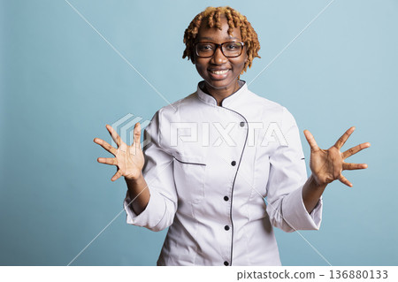 Smiling female chef in uniform gestures with open hands while standing in blue background studio. Professional cook expresses friendliness and enthusiasm, creating welcoming and positive culinary vibe Smiling female chef in uniform gestures with open hands while standing in blue background studio. Professional cook expresses friendliness and enthusiasm, creating welcoming and positive culinary vibe 136880133