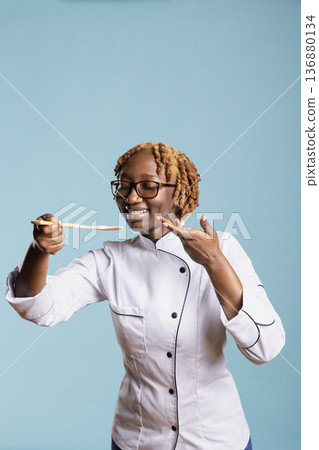 Joyful female chef in uniform tasting food using wooden spoon against blue background. Professional cook expressing satisfaction and creativity, showing passion and excellence for culinary arts. 136880134