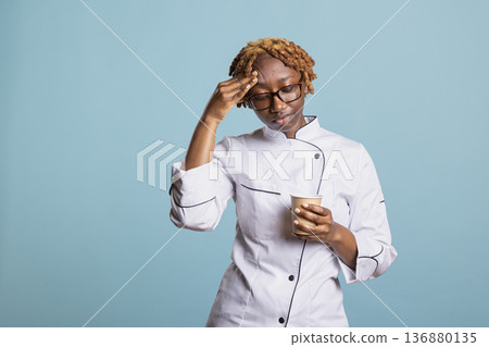 Exhausted female chef with glasses rubs forehead while holding disposable coffee cup after long day in kitchen. Tired restaurant staff member takes short break, needing caffeine to recharge and relax. Exhausted female chef with glasses rubs forehead while holding disposable coffee cup after long day in kitchen. Tired restaurant staff member takes short break, needing caffeine to recharge and relax. 136880135