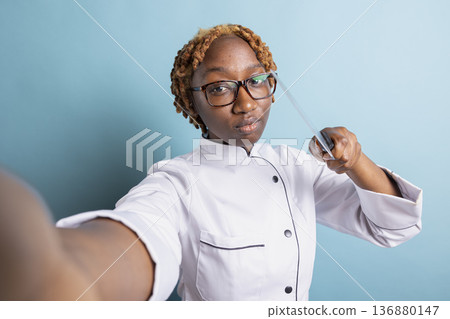 African American professional cook playfully pointing kitchen knife toward camera, recording culinary video. Confident female chef shows seriousness in preparation for gourmet challenge. African American professional cook playfully pointing kitchen knife toward camera, recording culinary video. Confident female chef shows seriousness in preparation for gourmet challenge. 136880147