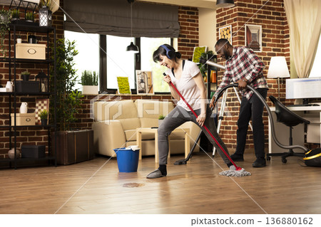 Cheerful housewife enjoys music while cleaning, playfully performing mini concert with mop stick as microphone. Young woman sings favorite song from headphones, taking fun break from mopping floor. Cheerful housewife enjoys music while cleaning, playfully performing mini concert with mop stick as microphone. Young woman sings favorite song from headphones, taking fun break from mopping floor. 136880162