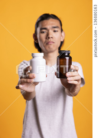Asian man holding two different pill bottles close to camera, showing various supplements. Unsure male individual extending arms while presenting vitamin or medication containers. 136880163