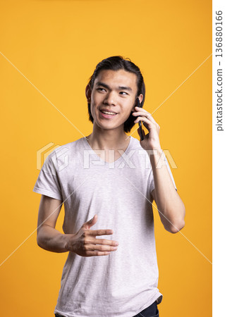Male freelancer with phone to ear, having pleasant conversation with colleagues, discussing business ideas. Asian man communicating happily with clients, friends or relatives using mobile device. Male freelancer with phone to ear, having pleasant conversation with colleagues, discussing business ideas. Asian man communicating happily with clients, friends or relatives using mobile device. 136880166