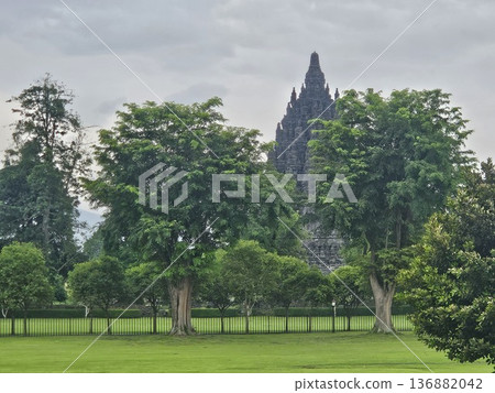 A majestic view of the ancient Prambanan Hindu temple towering behind lush green trees and a wide grassy field under a cloudy sky, showcasing Indonesia's historic stone architecture and heritage site. 136882042