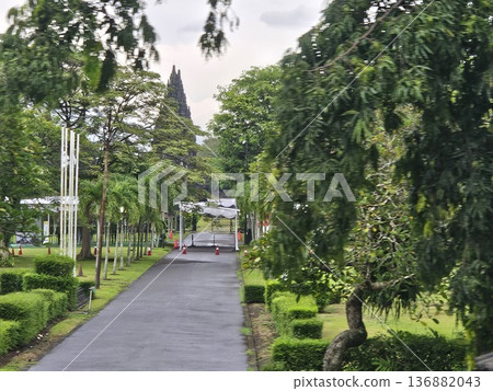 A majestic view of the ancient Prambanan Hindu temple towering behind lush green trees and a wide grassy field under a cloudy sky, showcasing Indonesia's historic stone architecture and heritage site. 136882043
