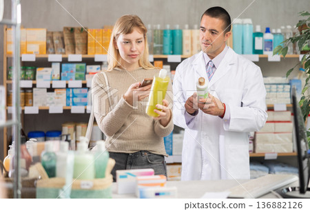Young woman in pharmacy scans QR code on hair loss shampoo, man pharmacist in background 136882126