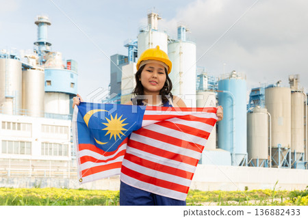 Happy asian girl in work clothes and hardhat with flag of malaysia standing in front of industrial scenery 136882433