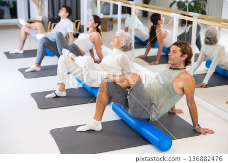 Positive athletic male performing set of exercises with pilates roller during group class in fitness studio. 136882476