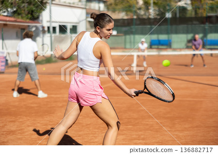 Attractive young girl in sportswear holding tennis racquet during ball serve. Family doubles match at tennis club Attractive young girl in sportswear holding tennis racquet during ball serve. Family doubles match at tennis club 136882717