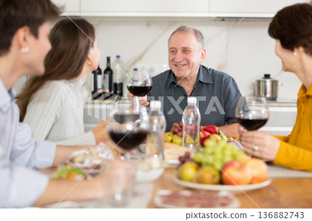 Senior man at kitchen table with wife, grown son and daughter, enjoying conversation at festive table with wine and snacks 136882743