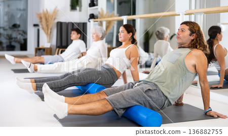 Positive athletic male performing set of exercises with pilates roller during group class in fitness studio. 136882755