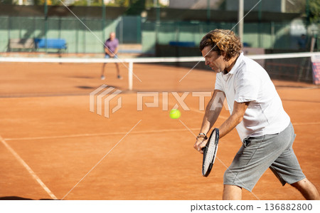 Handsome athletic man serving with racket in hands while practicing on tennis court in day 136882800