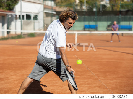 Back view of active male player hitting ball with racket on tennis court on blurred background of opponents Back view of active male player hitting ball with racket on tennis court on blurred background of opponents 136882841