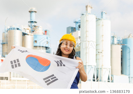 Happy young woman in helmet holding flag of South Korea against background of factory 136883170