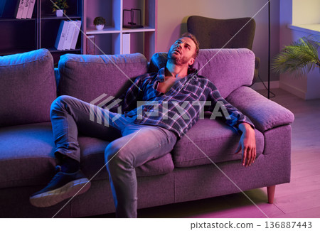 Exhausted man on couch under neon lights, evoking drugs, euphoria, and depression Exhausted man on couch under neon lights, evoking drugs, euphoria, and depression 136887443