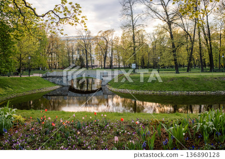 Beautiful Park Landscape With a Bridge Over a Calm River Surrounded by Vibrant Flowers and Lush Trees During Early Morning Light 136890128