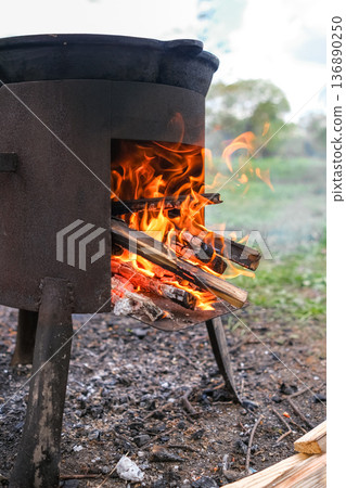 Fire Burns Brightly in a Rustic Metal Stove While Wood is Added for Cooking Outdoors in a Natural Setting During Daytime 136890250