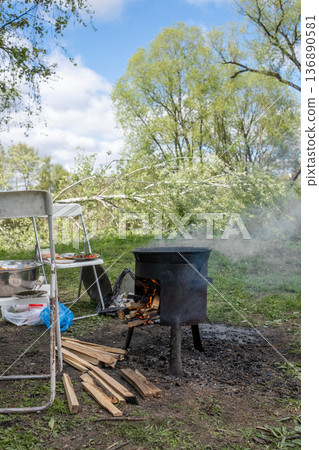 Cooking Outdoors With a Pot Over an Open Fire in a Lush Green Setting Surrounded by Trees and a Few Scattered Chairs Cooking Outdoors With a Pot Over an Open Fire in a Lush Green Setting Surrounded by Trees and a Few Scattered Chairs 136890581