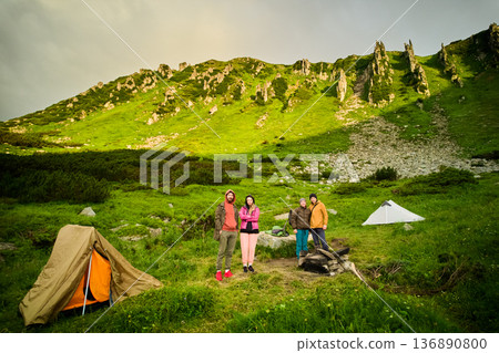 Group of happy campers stands near white tent in lush, green mountain setting. Rocky cliffs and vibrant vegetation, creating a stunning and adventurous atmosphere. 136890800