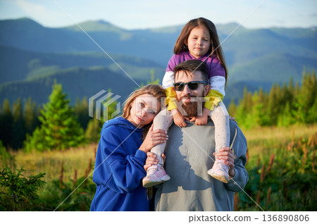 Family stands together in scenic meadow with backdrop of rolling mountains. Father carries his daughter on shoulders, both smiling and enjoying view, while mother stands beside them. Family stands together in scenic meadow with backdrop of rolling mountains. Father carries his daughter on shoulders, both smiling and enjoying view, while mother stands beside them. 136890806