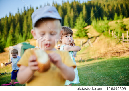 Young girl looks over her shoulder while holding bun. Boy in yellow shirt and cap focuses on his snack in blurred foreground. Another child plays, surrounded by pine trees and rolling hills. Young girl looks over her shoulder while holding bun. Boy in yellow shirt and cap focuses on his snack in blurred foreground. Another child plays, surrounded by pine trees and rolling hills. 136890809