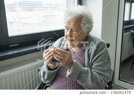 Senior woman in wheelchair holding warm cup and looking out of window. 136891479