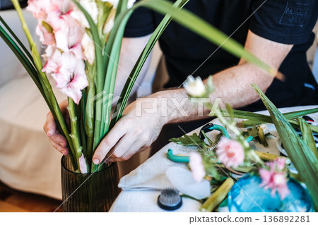 Man arranges gladiolus flowers in glass vase on white table. Men in floristry, breaking gender norms, male florist, creative masculinity, gender-neutral hobbies. 136892281