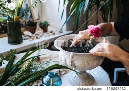Hands secure wire mesh in stone urn next to pink aster in blue glass vase. artisan floral design, handmade ceramic vases, craft focused floristry, diy flower frog arrangements. 136892282