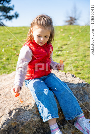 Little girl sitting on decorative stones blowing soap bubbles in city park. Spring childhood leisure and joyful outdoor play. 136893412