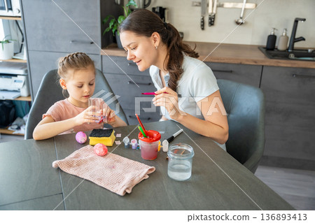 Mother and daughter decorating Easter eggs with colorful stickers while sitting at table at home. Family Easter craft activity, shared creativity and joyful holiday preparation indoors. 136893413