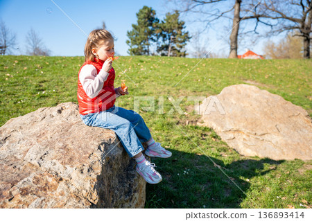 Little girl sitting on decorative stones blowing soap bubbles in city park. Spring childhood leisure and joyful outdoor play. 136893414