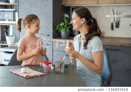Child painting bright designs on Easter egg while sitting at table. The image reflects creativity and celebration of spring rituals. 136893428
