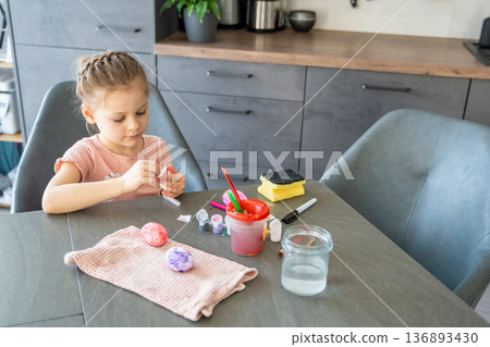 Little girl decorating Easter eggs with colorful stickers while sitting at table at home. Creative Easter activity, simple egg decoration technique and joyful holiday preparation indoors. 136893430