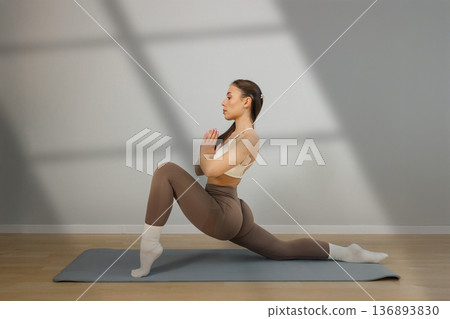 Young athletic woman practicing yoga lunge pose indoors. Female holding prayer hands while performing low lunge stretch on yoga mat, balance, flexibility and mindful workout in minimal studio Young athletic woman practicing yoga lunge pose indoors. Female holding prayer hands while performing low lunge stretch on yoga mat, balance, flexibility and mindful workout in minimal studio 136893830