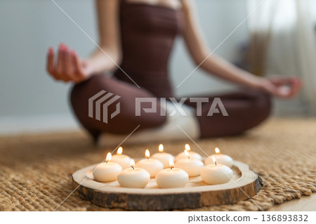 Woman practicing meditation indoors. Lotus pose, burning candles on wooden tray, calm atmosphere, mindfulness and wellness lifestyle concept. Woman practicing meditation indoors. Lotus pose, burning candles on wooden tray, calm atmosphere, mindfulness and wellness lifestyle concept. 136893832