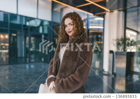 Young woman posing near modern glass building in the city. Stylish casual outfit, confident look, contemporary urban lifestyle, evening light and fashionable atmosphere. Young woman posing near modern glass building in the city. Stylish casual outfit, confident look, contemporary urban lifestyle, evening light and fashionable atmosphere. 136893833