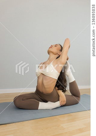 Young flexible woman practicing advanced yoga stretch indoors. Female performing deep backbend pose with leg hold on yoga mat, flexibility training, strength and balance in minimal studio. 136893838