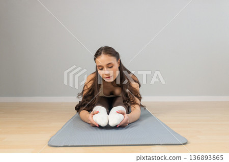 Young woman stretching legs during yoga practice indoors. Forward fold seated pose on mat, flexible athletic body, calm focused expression, minimal studio interior, soft natural light Young woman stretching legs during yoga practice indoors. Forward fold seated pose on mat, flexible athletic body, calm focused expression, minimal studio interior, soft natural light 136893865