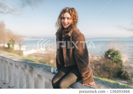 Young woman sitting on seaside promenade railing, enjoying sunny day by the sea. Casual outfit, relaxed pose, coastal city lifestyle, freedom and calm outdoor atmosphere. 136893866
