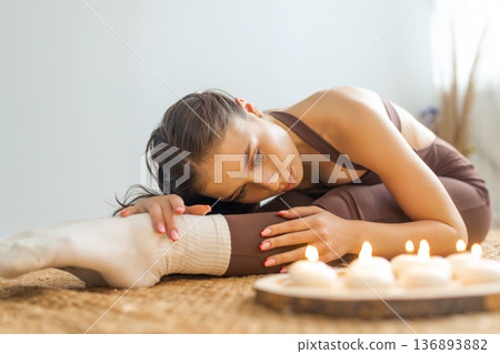 Young woman practicing yoga stretching indoors. Forward bend pose on natural mat, soft candlelight, calm minimal interior, relaxation, mindfulness and wellness lifestyle concept. 136893882
